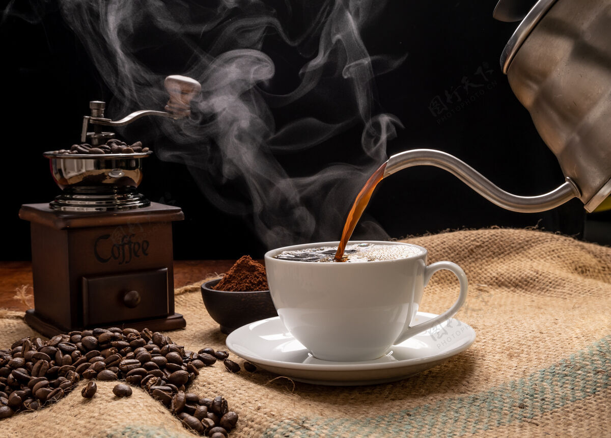 咖啡蒸汽咖啡杯与研磨机 烤啤酒 咖啡粉和水壶在粗麻布黑森麻布在垃圾木桌子的背景灯光研磨机地面