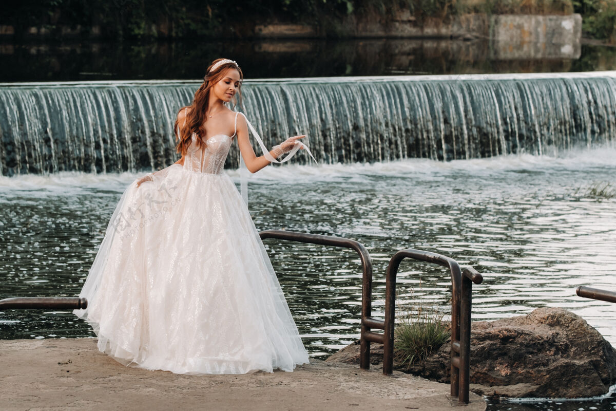 优雅一位穿着白色礼服 戴着手套的优雅新娘站在公园的河边 享受着大自然婚纱男人自然