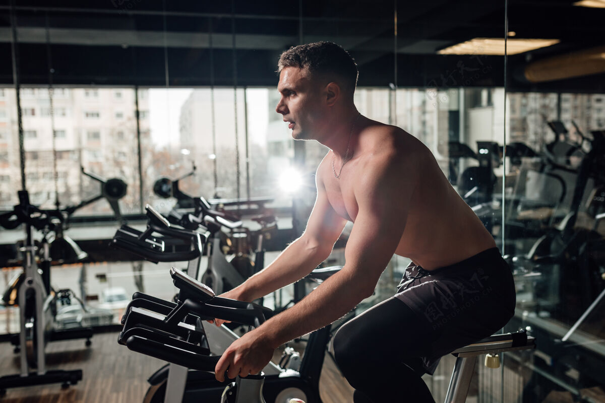 物理侧视图赤膊上阵的年轻人在自行车上锻炼在镜子附近的现代健身房强壮动力自行车