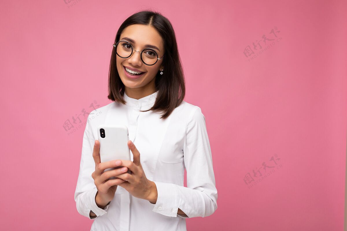互联网迷人的微笑的年轻黑发女子身穿白色上衣和眼镜站在粉红色的墙上使用手机短信孤立电话移动手机