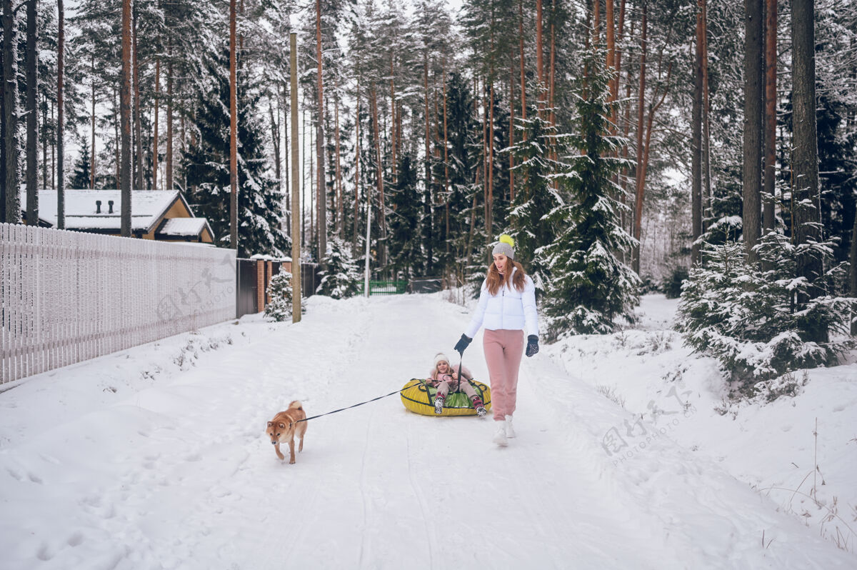 休闲快乐的妈妈和可爱的小女孩穿着粉色温暖的外套 在户外的雪白寒冷的森林里 玩着充气雪地管和红色的石坝犬寒冷家庭玩耍