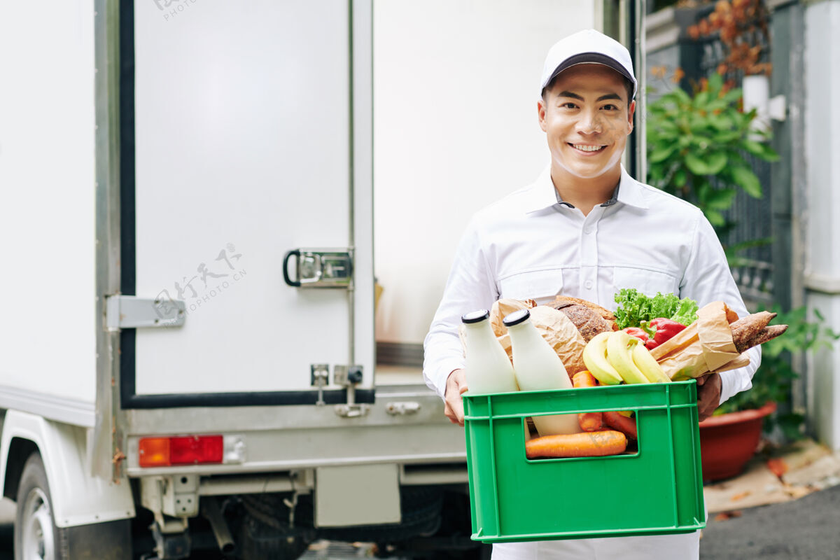 男人正面的越南年轻信使的肖像 拿着装有新鲜水果 面包和瓶装牛奶的塑料盒亚洲蔬菜工作服