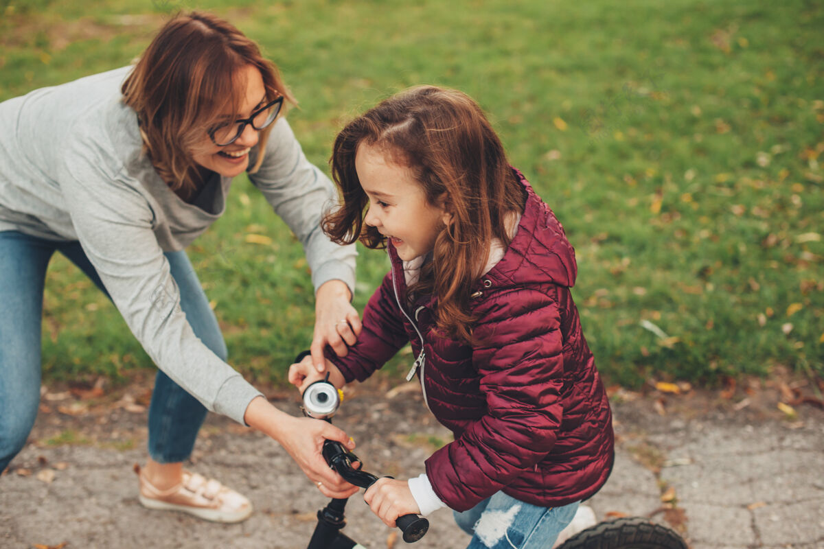 成人侧视图一个可爱的小女孩在自行车上笑 而她的母亲发现她在公园户外笑教学学习情感