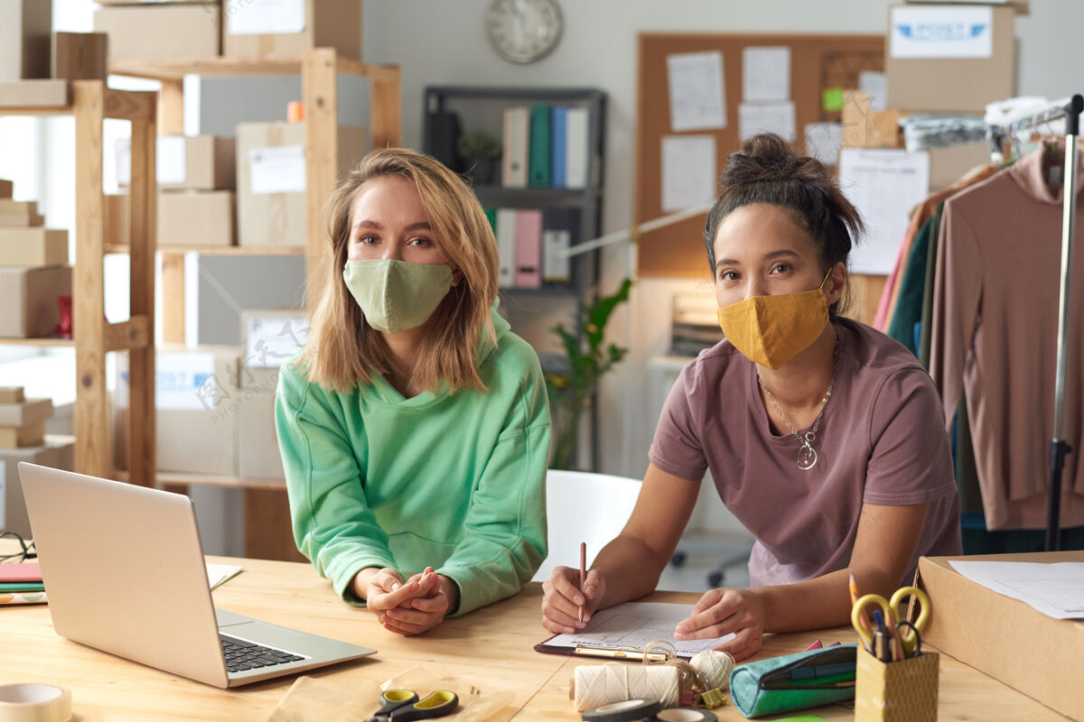 使用笔记本电脑车间里 两个戴着防护面具的年轻女子拿着笔记本电脑在桌边工作裁缝同事