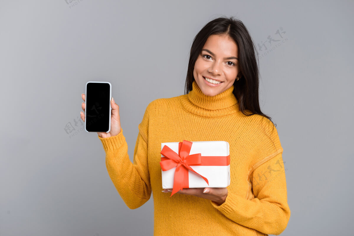 漂亮一个快乐随意的年轻女子的肖像被隔离在灰色的墙上 显示手机屏幕空白 手持礼品盒购物空白休闲