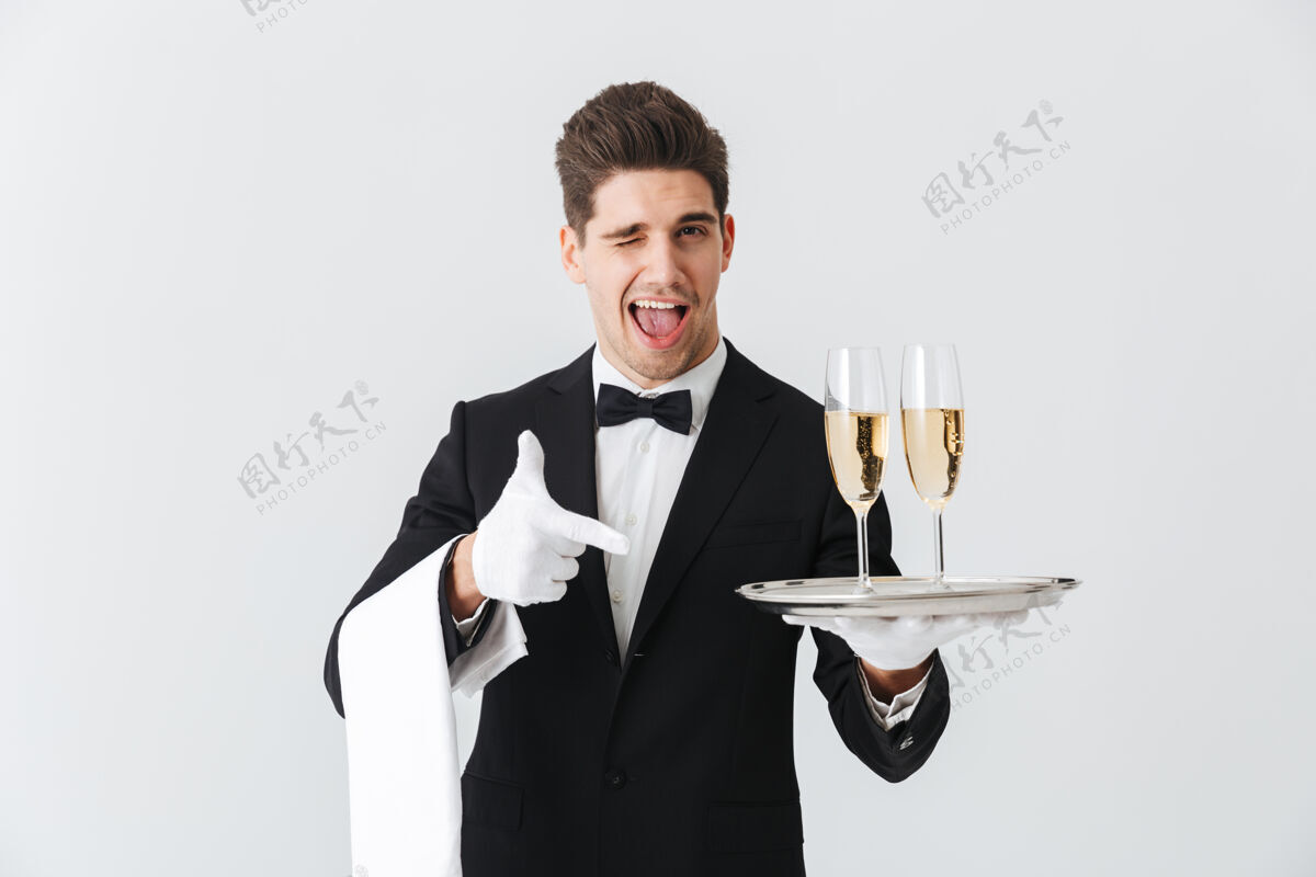 酒杯一个身着燕尾服的微笑的年轻侍者的画像在白墙上为你提供了一杯香槟男性服务饮料