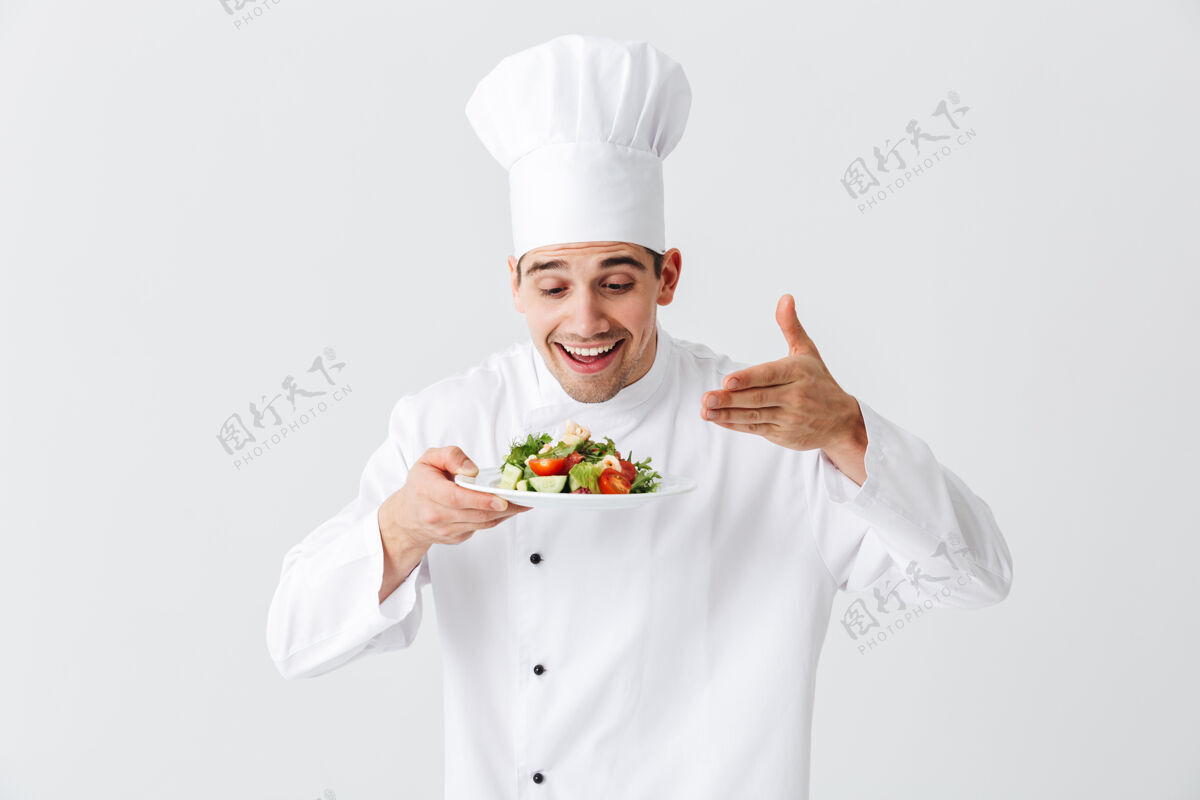 服务兴奋的男厨师穿着制服 把新鲜的蔬菜沙拉放在隔离在白色墙上的盘子上烹饪成人男性