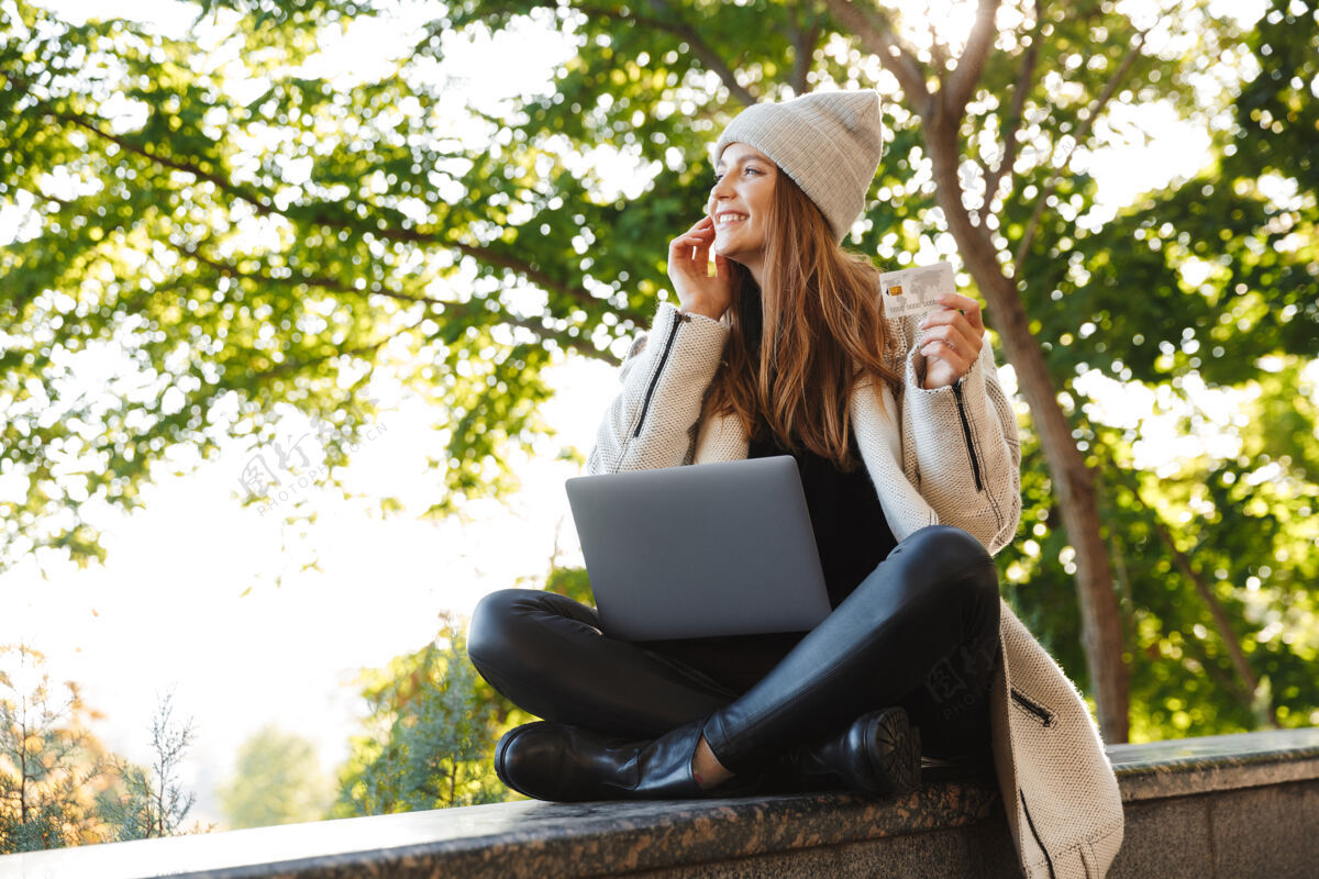 消费主义快乐的年轻女子穿着秋大衣 戴着秋帽坐在户外 用笔记本电脑 展示塑料信用卡成功客户在线