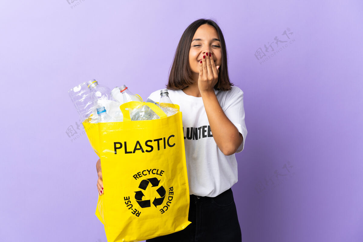 青年年轻的拉丁女子拿着一个装满废纸的回收袋来回收孤立的紫色 开心地笑着用手捏着圆锥形的嘴生态表达封面