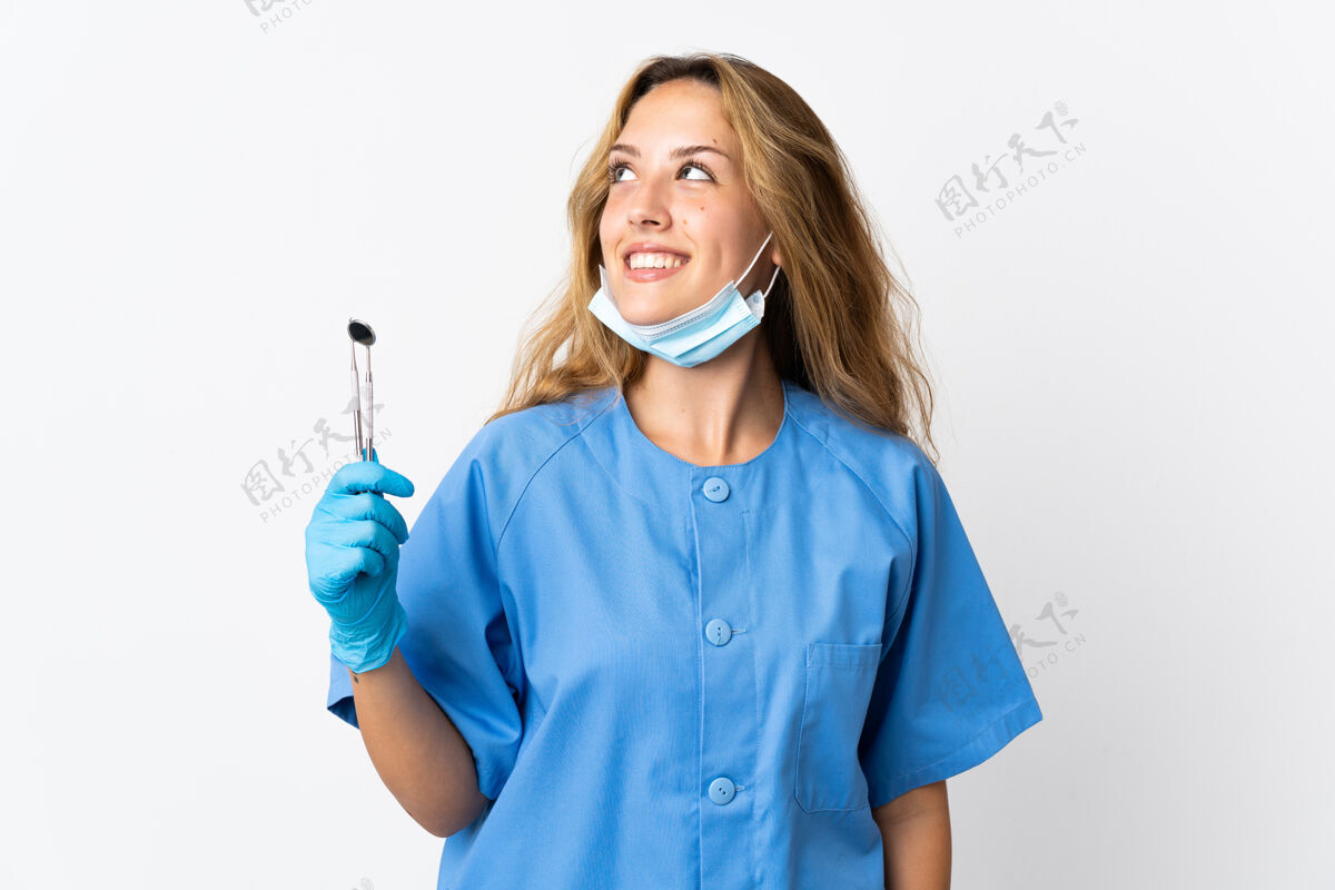 成年人女牙医拿着工具孤立地站在白纸上一边想一边抬头看健康人女人