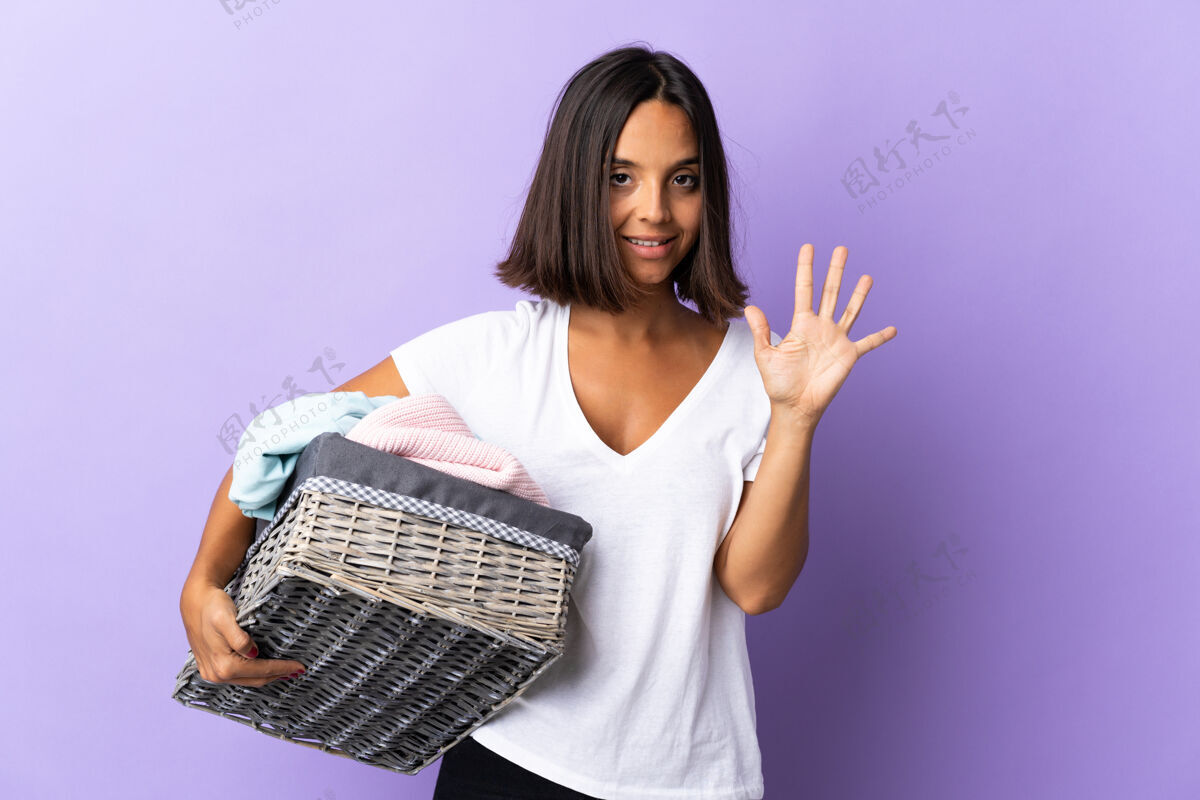 计数年轻的拉丁女子拿着一个衣篮 在紫色的地上用手指数着五个脏青年篮子