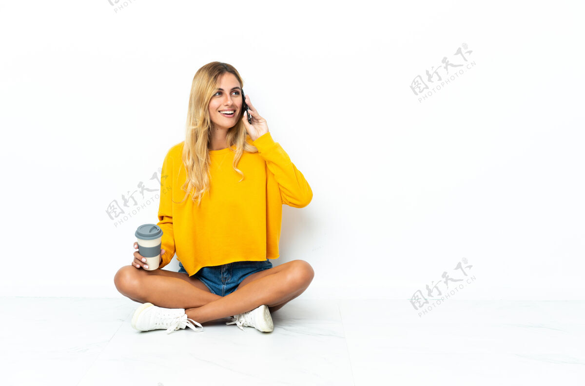 地板年轻的金发乌拉圭女子坐在地板上 隔着白色的咖啡拿着带走的手机房间全身成人