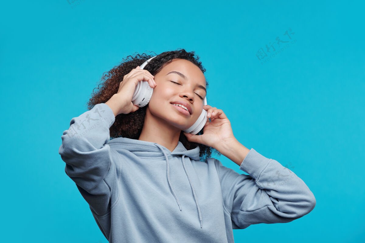 享受内容迷人的非洲裔美国女孩在无线耳机保持眼睛紧闭 同时享受喜爱的歌曲 蓝色背景女人欢呼美丽