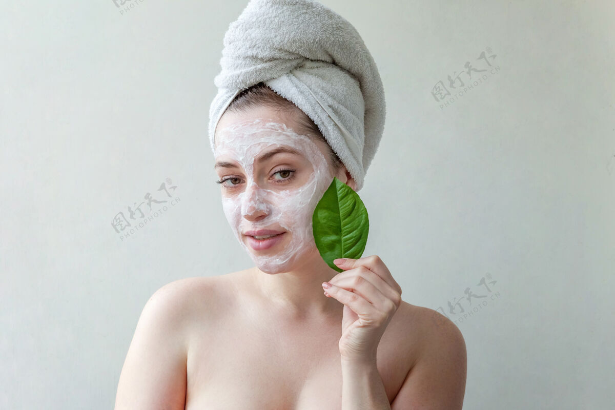 美容头戴毛巾 脸上涂有白色滋养面膜或乳霜 手拿绿叶 白色背景的美女肖像美容师营养面部