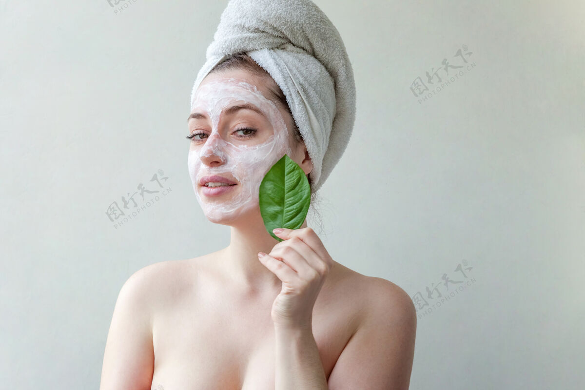 健康头戴毛巾 脸上涂有白色滋养面膜或乳霜 手拿绿叶 白色背景的美女肖像皮肤科女孩皮肤护理