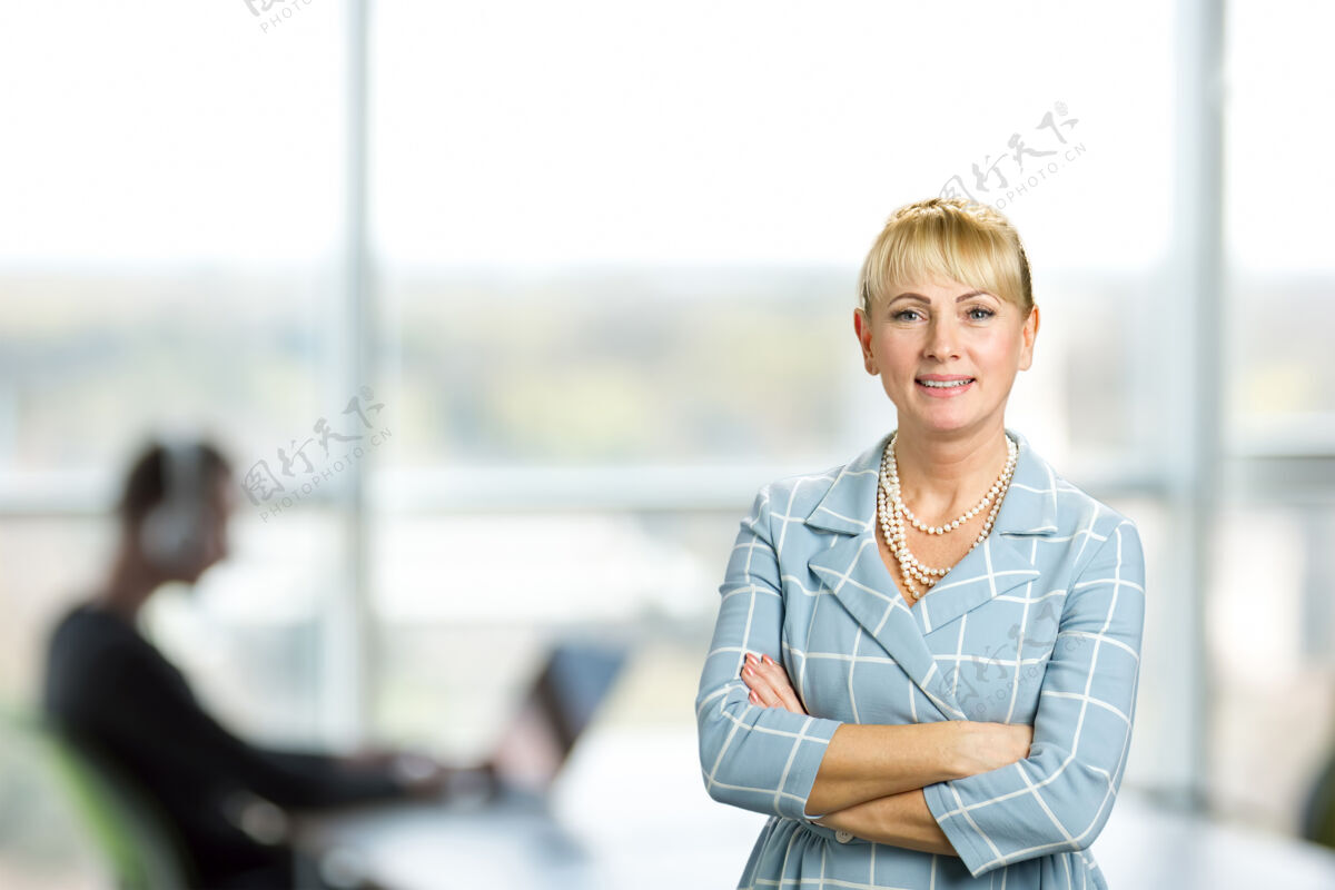 漂亮微笑的女人交叉双臂成熟的女人交叉双臂站在办公室的窗户上肖像中年女人
