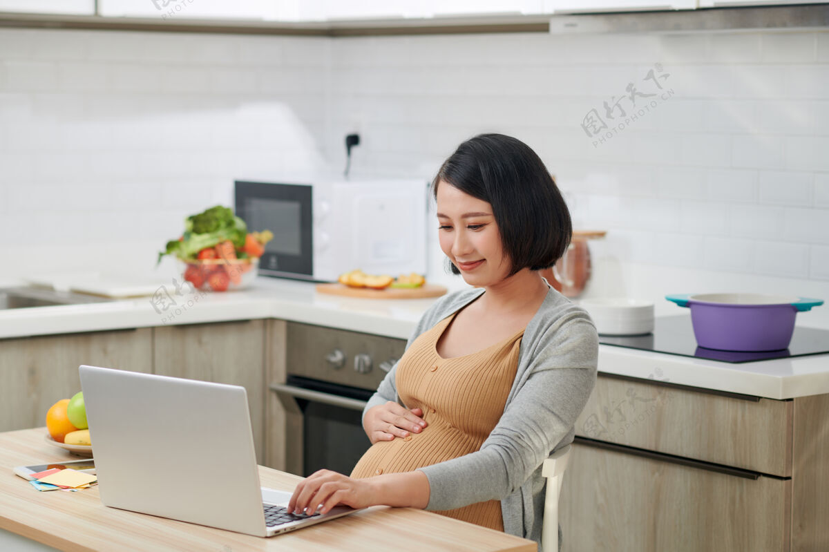 年轻人侧视图有吸引力和进步的年轻妈妈坐在椅子后面的桌子里面明亮的单位和使用pc笔记本电脑早餐室内厨房