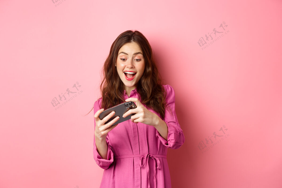 快乐科技概念漂亮的现代女孩在智能手机上玩赛车电子游戏 轻触手机屏幕 微笑着快乐 粉色的墙壁积极购物移动