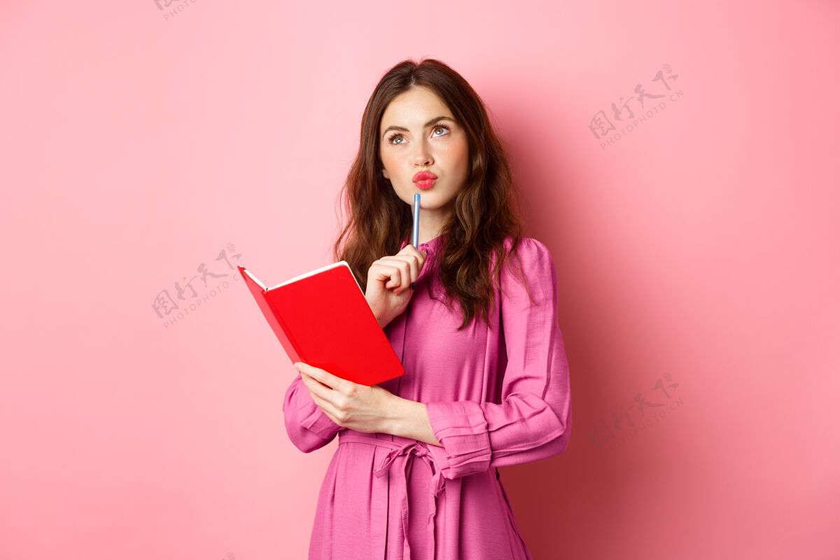 惊人漂亮的年轻女子看起来很体贴 写在笔记本上 拿着计划者或日记 计划她的日程 站在粉红色的墙上候选人女人微笑