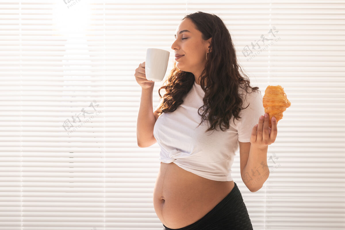 怀孕早上吃早餐时 美丽的孕妇手里拿着牛角包和一杯咖啡胃黑发责任