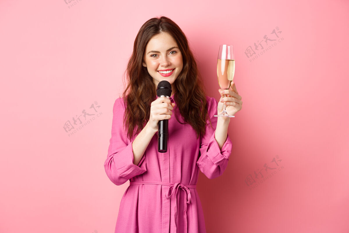 演讲庆祝和节日的概念美丽的年轻女子说祝酒词 提高杯chamapne同时发言 手持麦克风 站在粉红色的墙壁复制空间快乐兴奋微笑