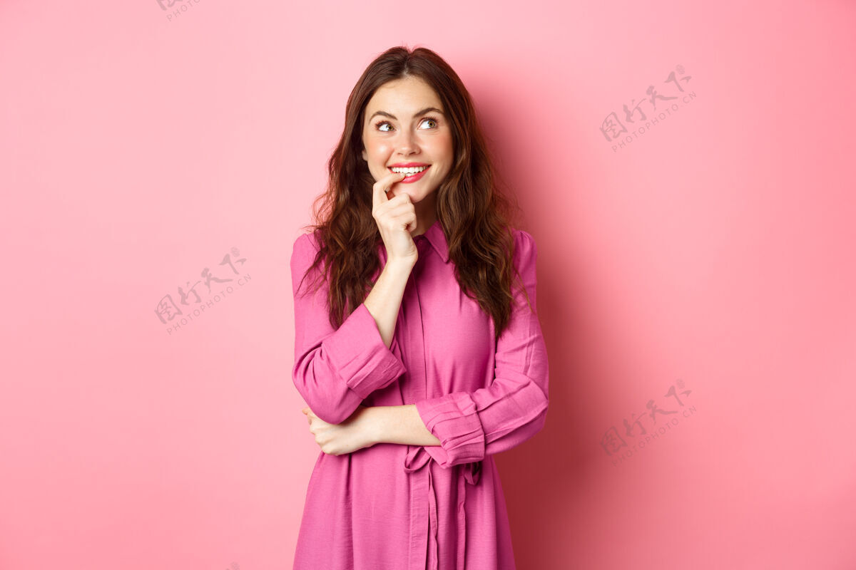 积极风骚的年轻女子站在粉色的墙上 一边若有所思 一边微笑 一边咬着手指思考 想象着什么微笑思想兴奋
