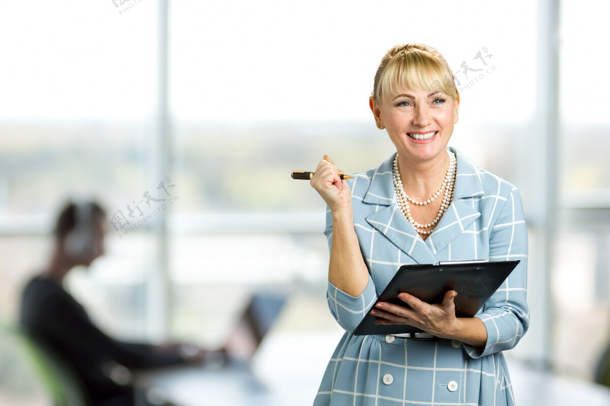 漂亮微笑的女人拿着文件夹积极成熟的女人拿着剪贴板和笔站在办公室的窗户上肖像笔记女性
