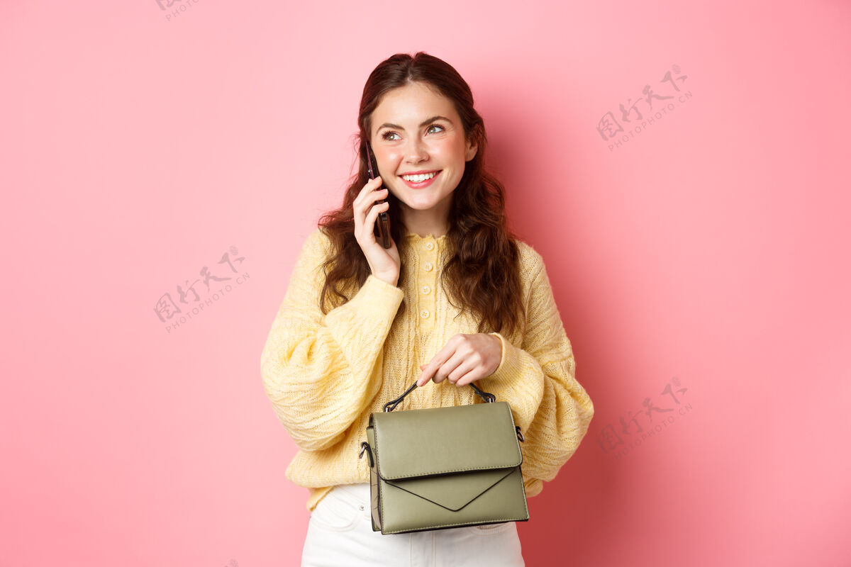 成人漂亮时尚的女士打电话 微笑着和别人打电话 拿着钱包 站在粉红色的墙上时尚黑发时尚
