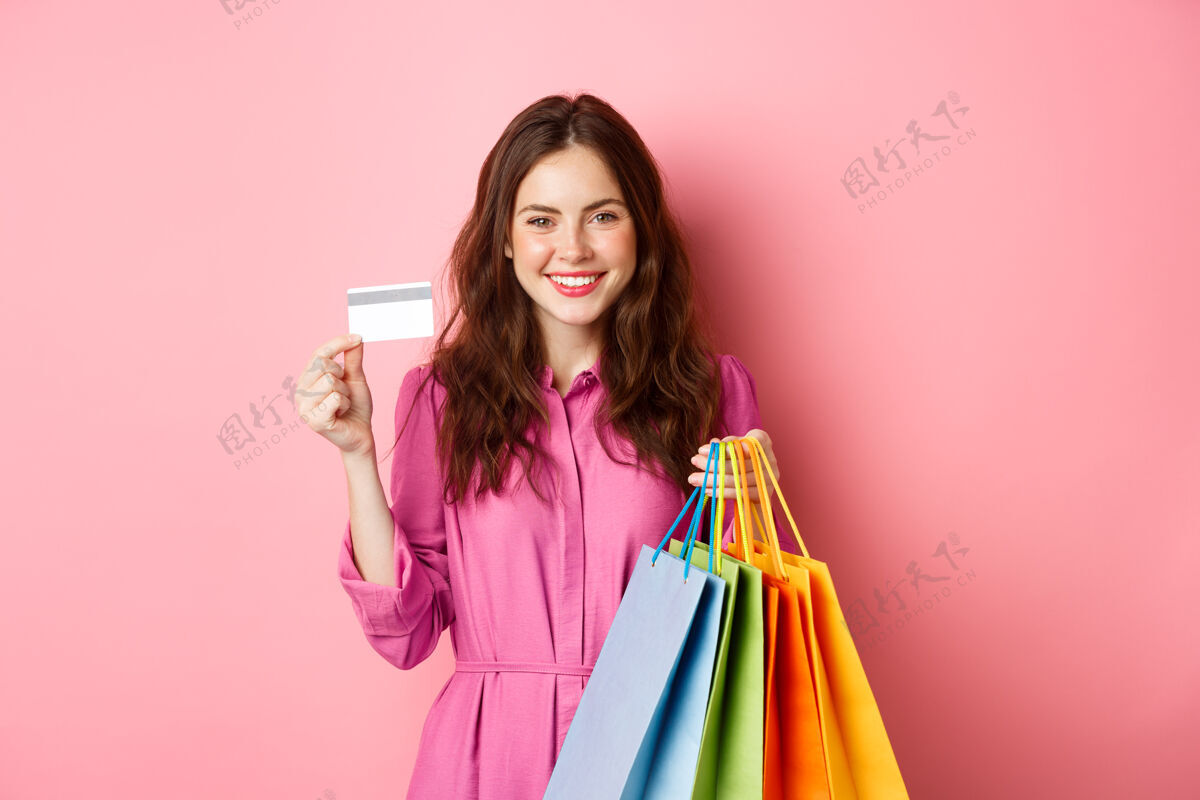 女人年轻的黑发女子拿着购物袋 展示塑料信用卡 面带微笑 站在粉红色的墙上信用卡候选人女性
