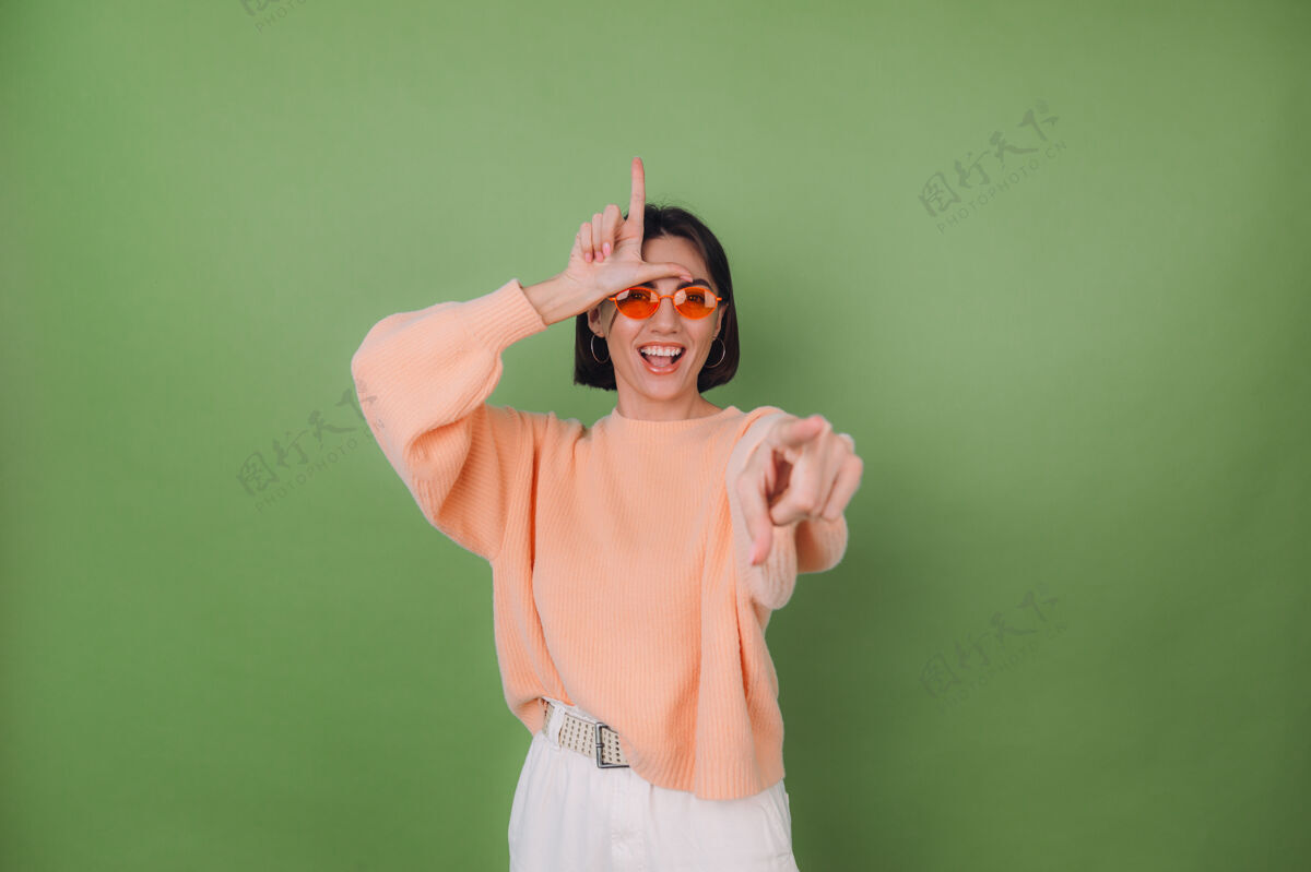 女性年轻女子穿着休闲桃色毛衣 戴着橙色眼镜 孤立地站在绿橄榄墙上 指着你嘲笑愚人节展示的号角符号复制空间手势角点