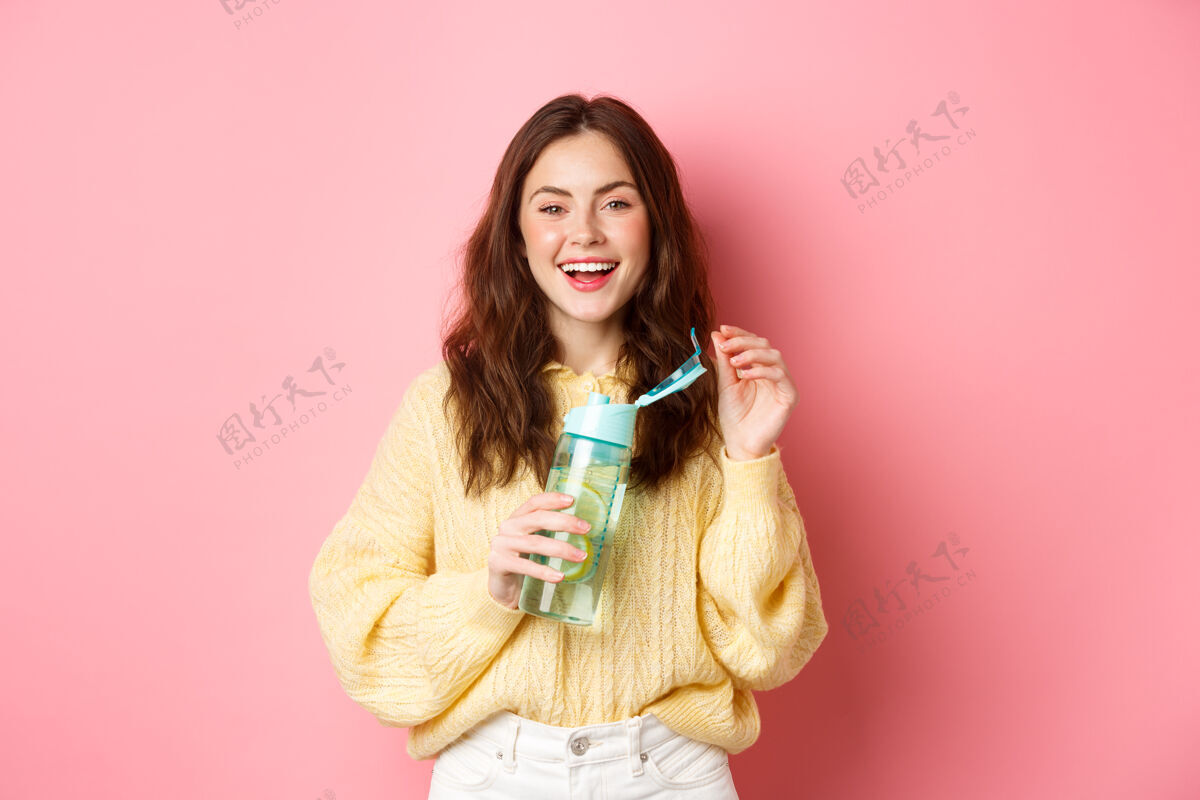 女人快乐的黑发女人的肖像 卷发 喝着柠檬水 笑着 感觉健康 站在粉红色的墙上女性乐观水瓶