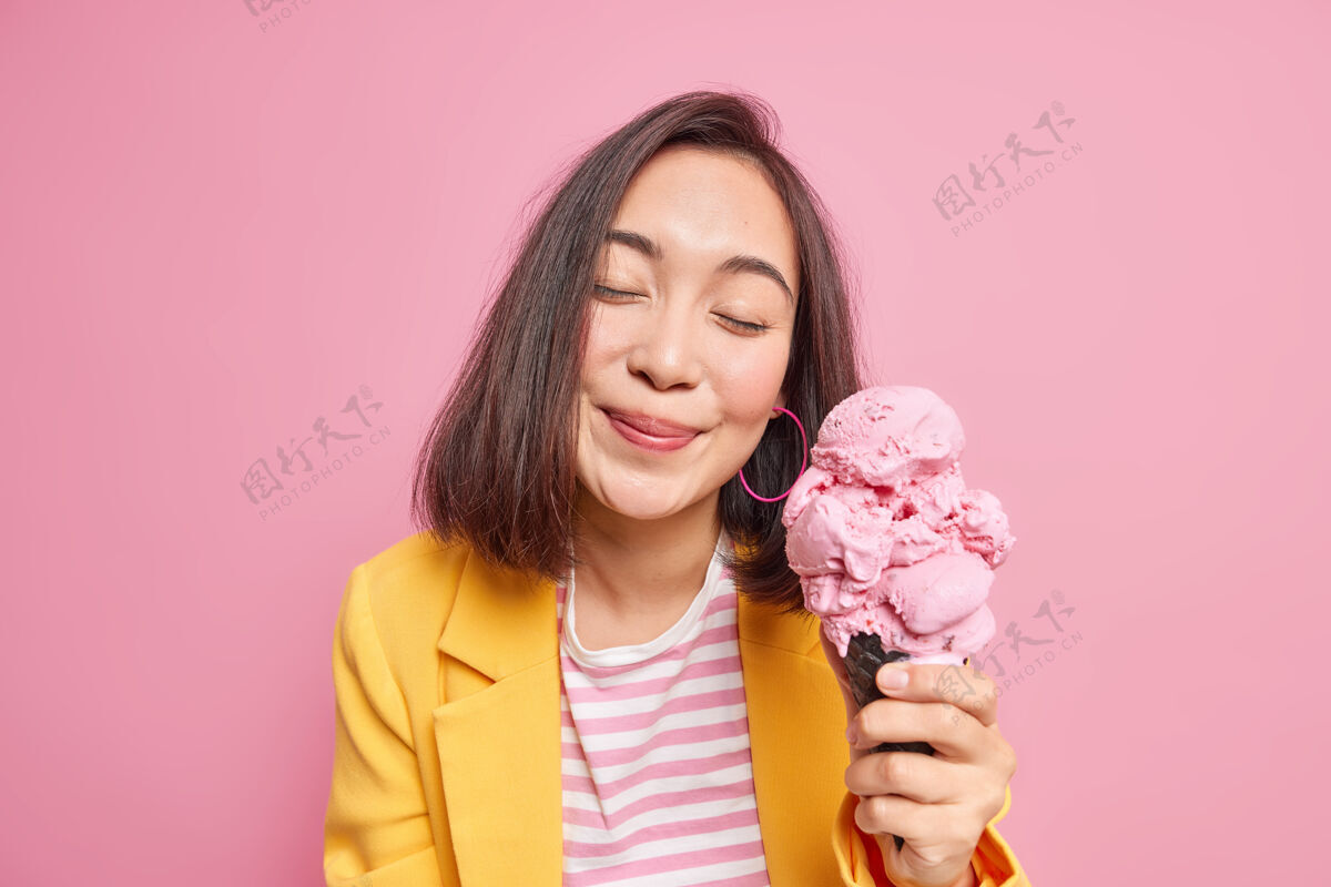 年轻可爱女人的室内镜头闭上眼睛看不到快乐 吃着美味的冰淇淋心情好穿着时髦的衣服美丽的东方女模特摆出美味的冷冻小吃欢呼微笑女性