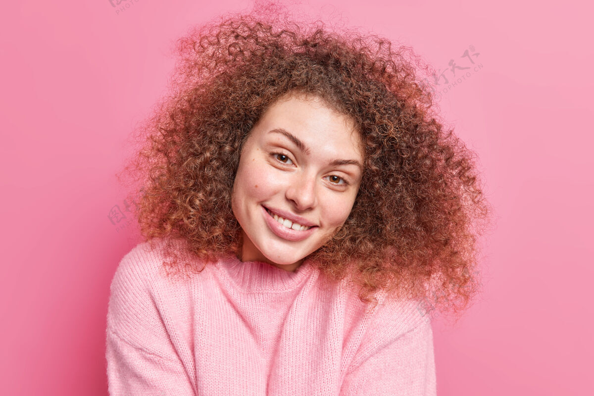 姿势正面诱人的女性肖像 卷曲浓密的头发 温柔的微笑 显示洁白的牙齿 健康的皮肤 穿着休闲套头衫 隔离在粉色的墙壁上自然美的概念肖像女人感觉