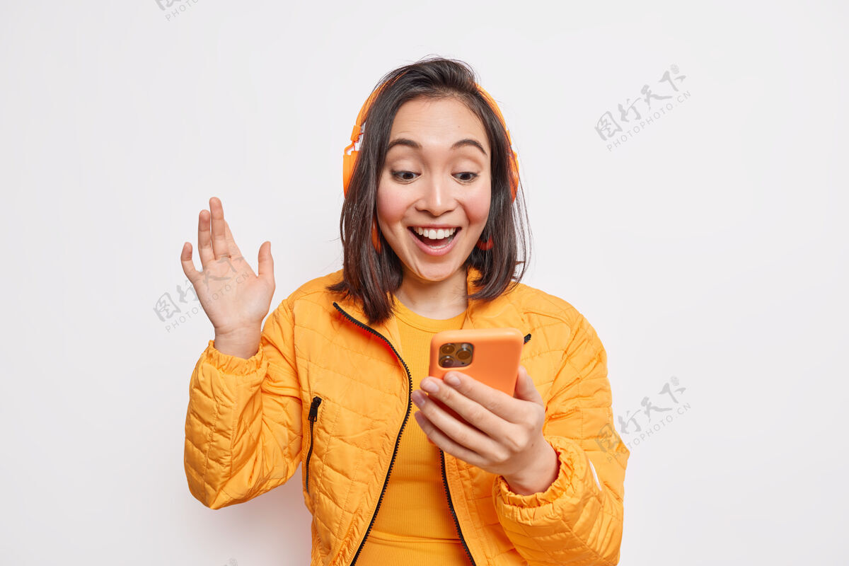 朋友开朗无忧无虑的亚洲少女手持智能手机挥手打视频电话在互联网音乐平台上挑选歌曲听最喜欢的播客用无线耳机穿着橙色夹克手亚洲人青少年