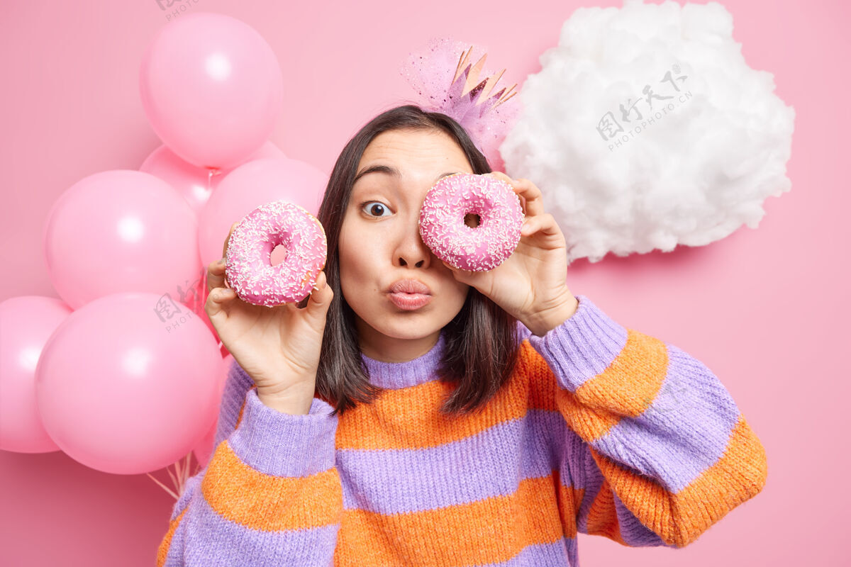 垃圾女人眼睛和美味的甜甜圈保持嘴唇合拢穿着休闲套头衫庆祝生日享受节日场合隔离在粉红色持有毛衣美味