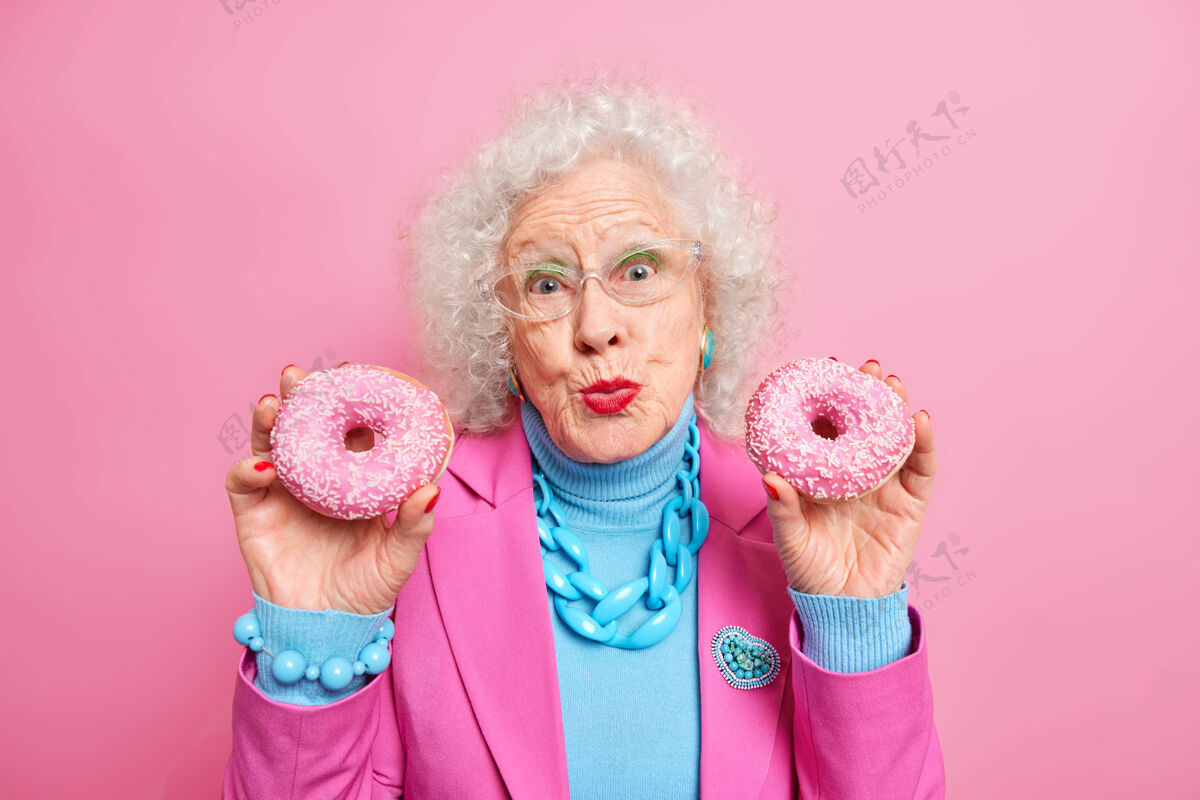 肖像可爱的高级女士拿着两个光滑的甜甜圈 双唇合拢 看起来很讨人喜欢 穿着时髦的服装 戴着项链和手镯饮食兴奋糖
