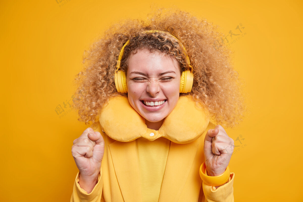音乐快乐积极向上的卷发女人脖子上戴着舒适的枕头减轻疼痛享受喜爱的音乐用无线耳机表达幸福隔着黄色的墙壁立体声歌曲庆典