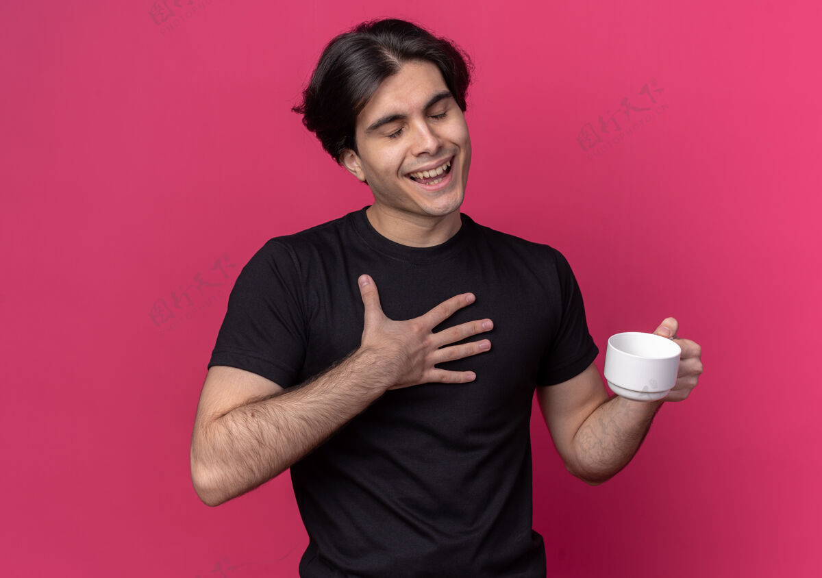 封闭喜欢闭上眼睛的年轻帅哥穿着黑色t恤 手里拿着一杯咖啡孤立在粉红色的墙上咖啡人姿势