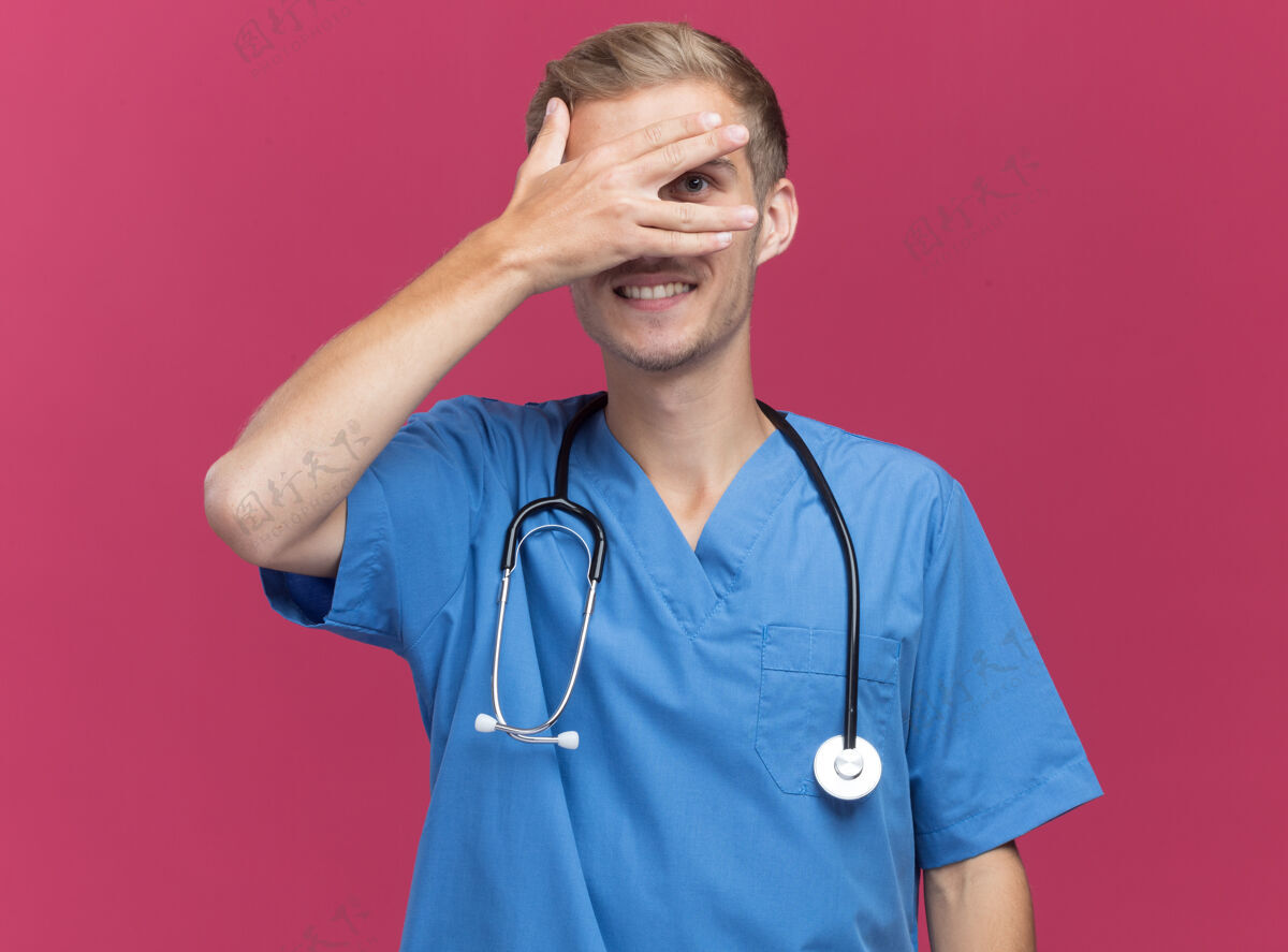人微笑的年轻男医生穿着医生制服 听诊器盖着脸 手被隔离在粉红色的墙上姿势穿着人