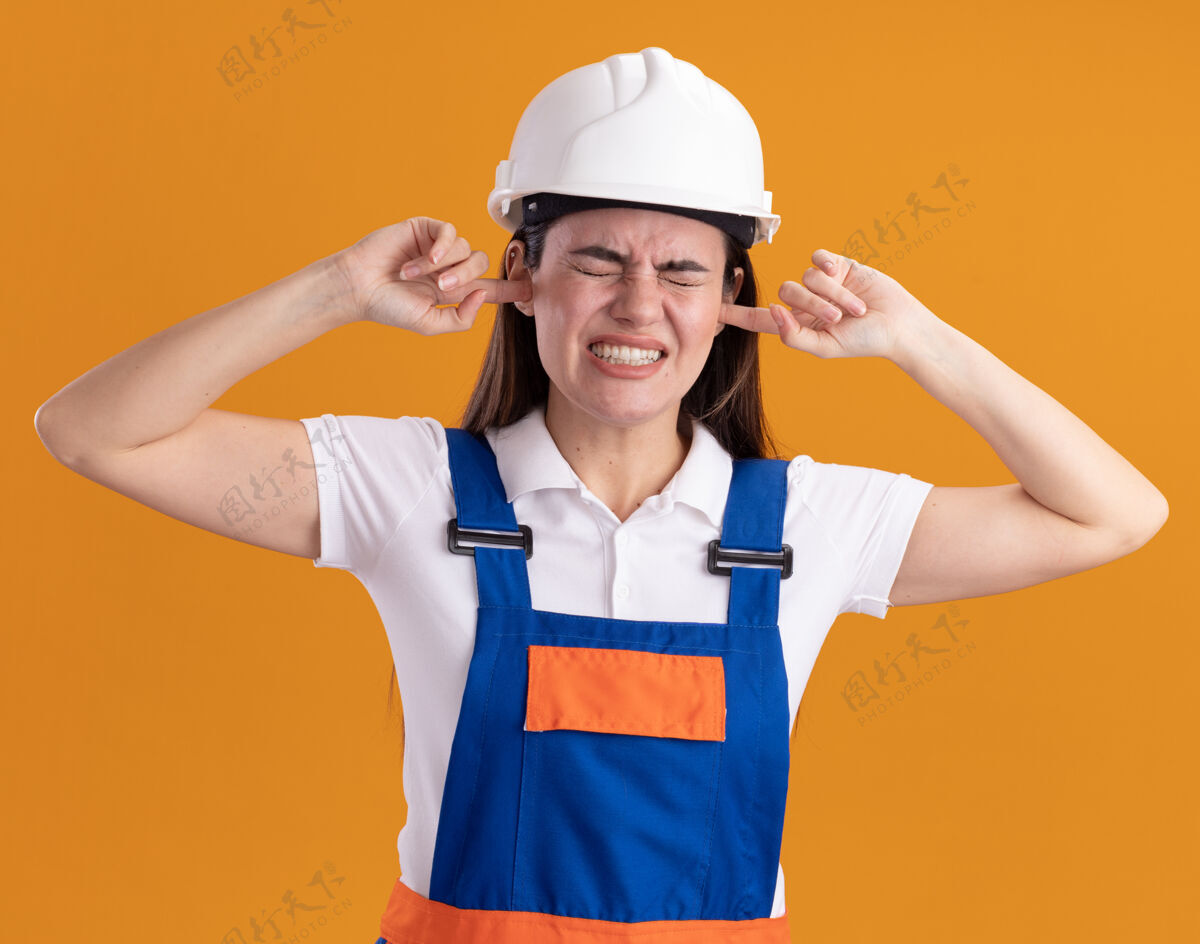 烦恼恼怒的年轻建筑妇女穿着制服 耳朵被隔离在橙色的墙上年轻人站建设者