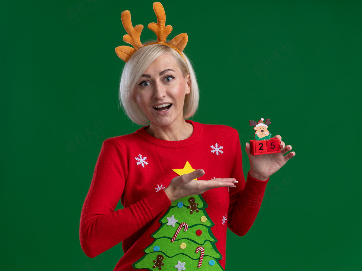 印象令人印象深刻的中年金发女人戴着圣诞驯鹿鹿角头带和圣诞毛衣拿着 用手指着圣诞驯鹿玩具和日期看起来孤立在绿色墙上驯鹿举行手