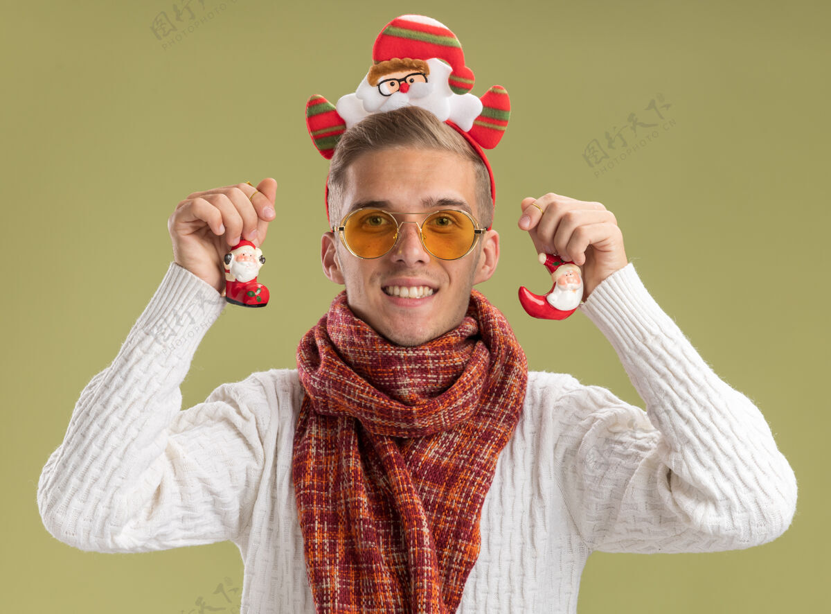 圣诞节微笑着的年轻帅哥戴着圣诞老人的头带和围巾看起来拿着圣诞老人的圣诞饰品孤立在橄榄绿的墙上头带帅哥年轻