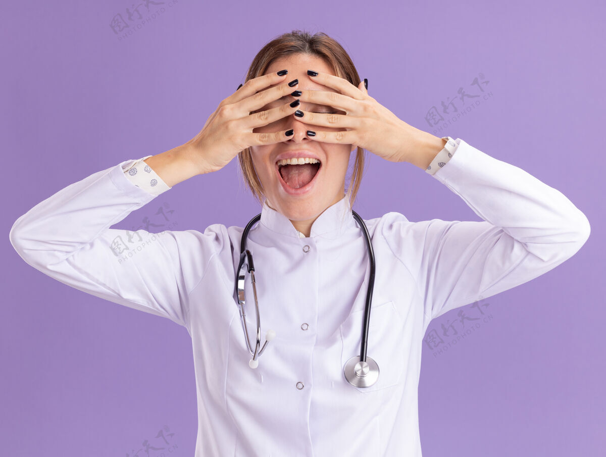 穿着兴奋的年轻女医生穿着医用长袍 听诊器捂着眼睛 手被隔离在紫色的墙上人脸兴奋