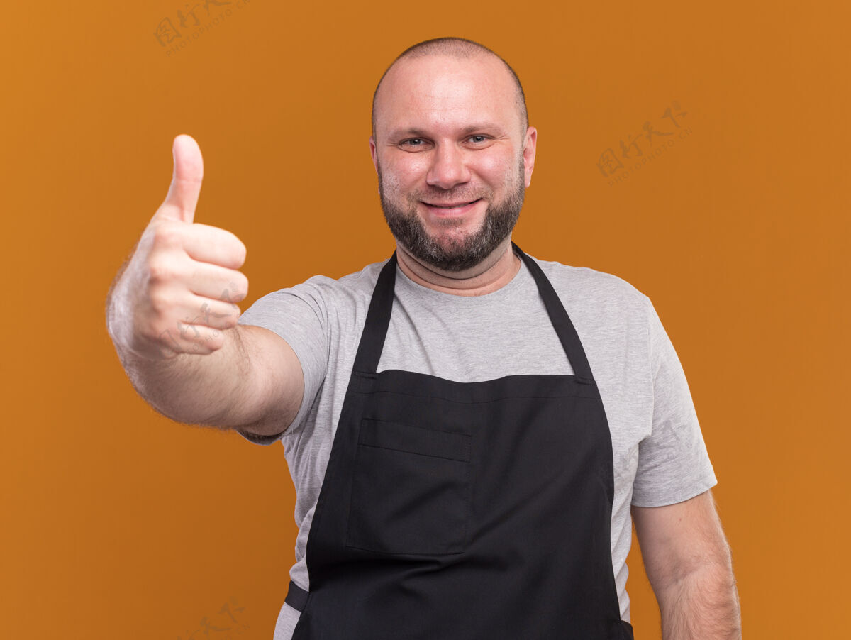 姿势微笑的中年男理发师穿着制服 在橙色的墙上孤立地竖起大拇指男性理发师公民