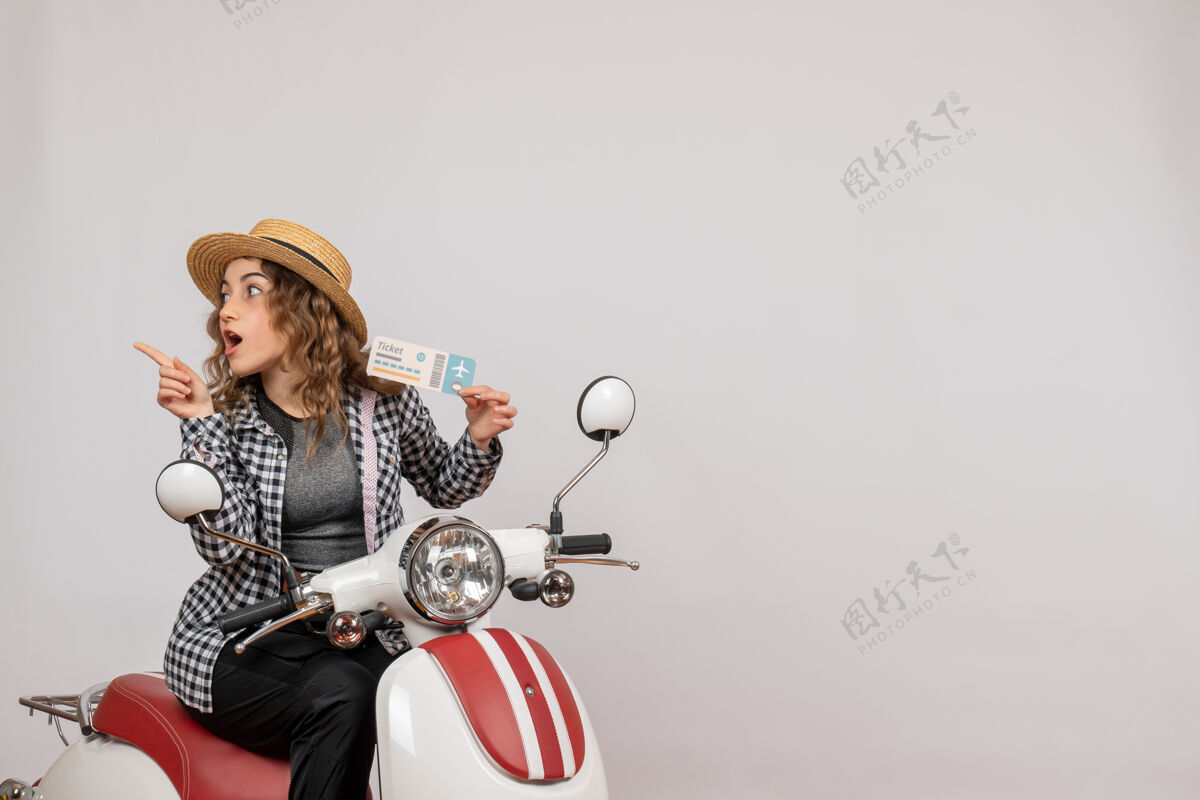 坐着前视图骑着轻便摩托车的年轻女孩举着车票指着左边灰色度假向上
