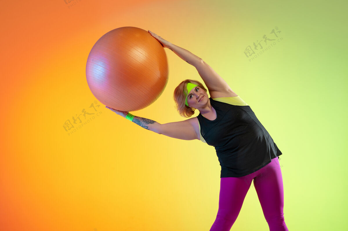 身体年轻的白人大号女模特在霓虹灯下的橙色渐变背景下训练用健身球做健身操运动理念 健康的生活方式 积极的身体 平等水平操作锻炼