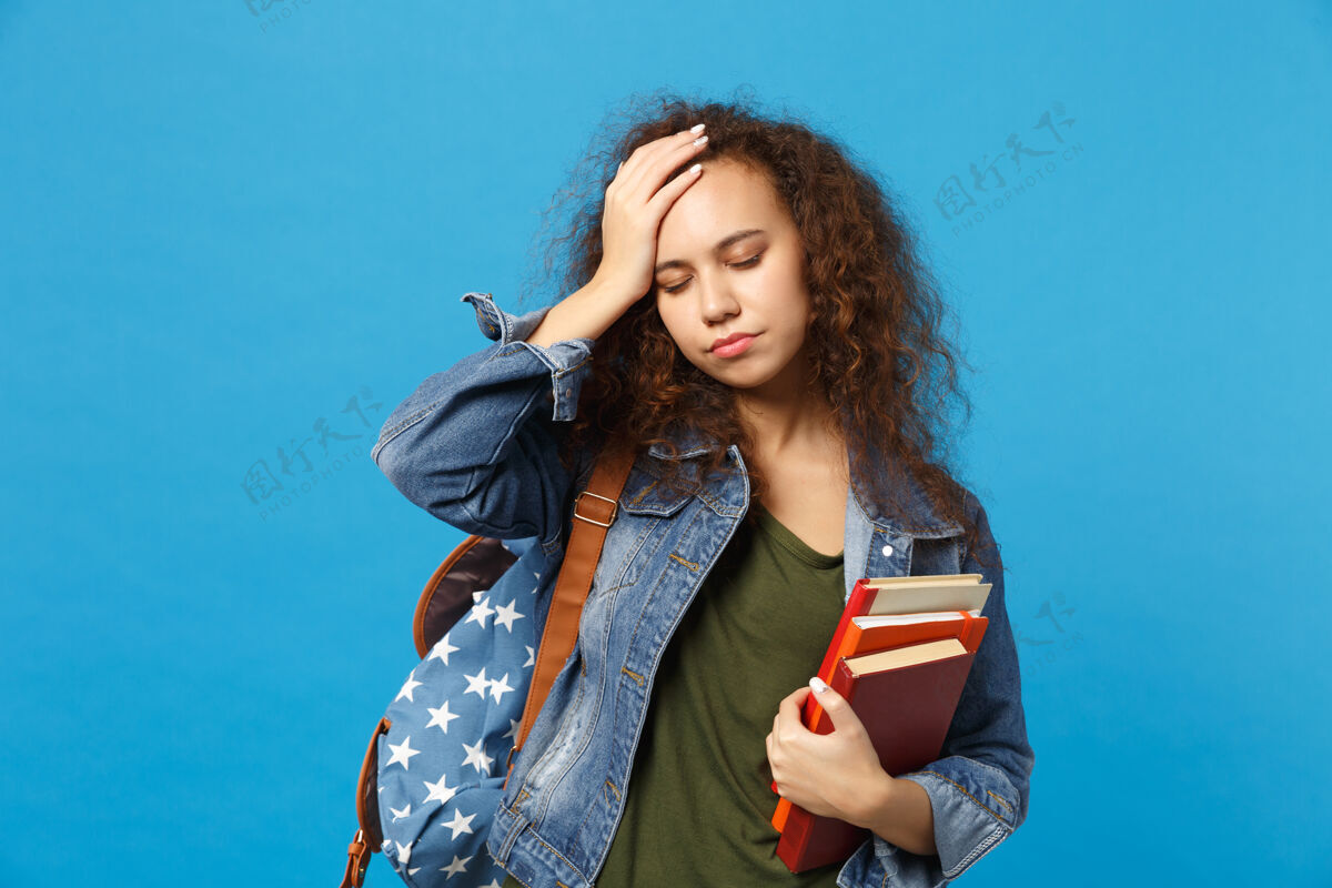 恼怒穿着牛仔服 背着书包的悲伤的年轻学生把书孤立地放在蓝色的墙上知识沮丧非洲