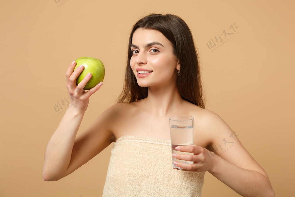 新鲜近距离拍摄完美肌肤的半裸女人 裸妆将苹果和水隔离在米色粉彩墙上手玻璃器皿水