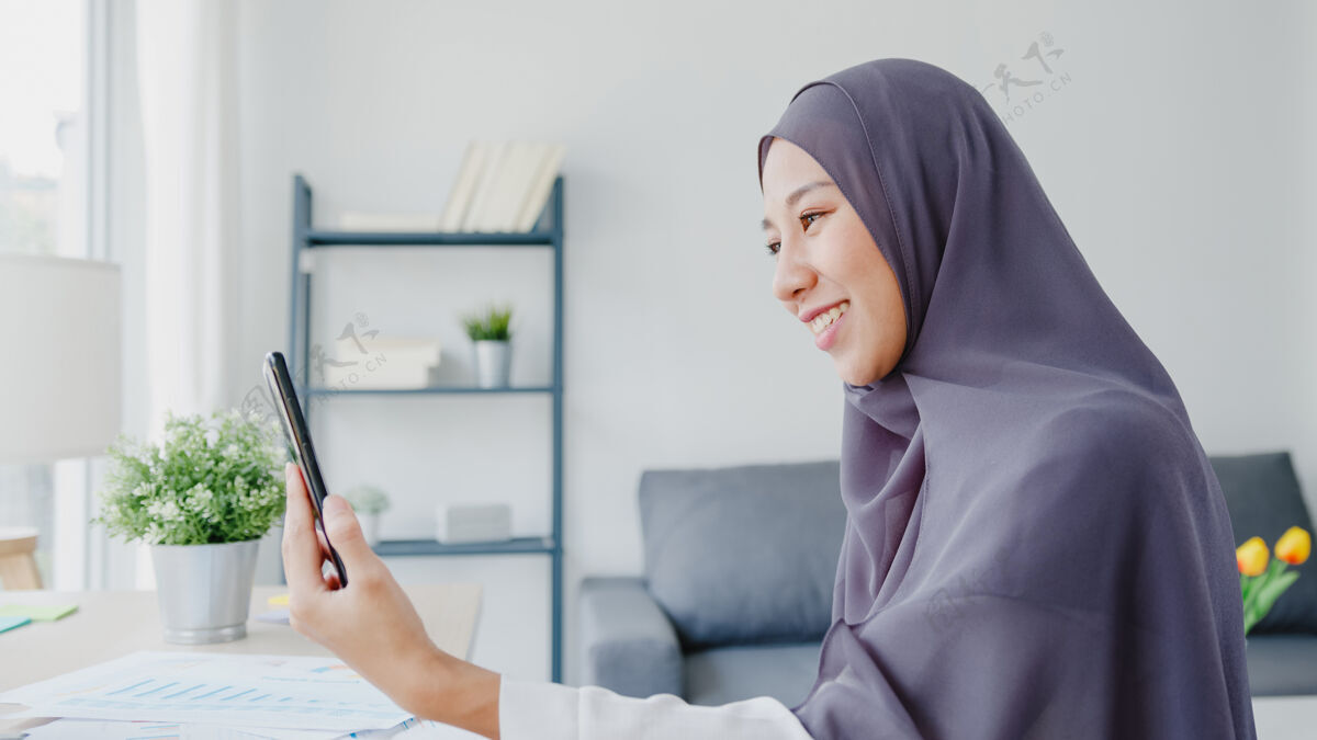 女性年轻的亚洲穆斯林女商人使用智能手机通过视频聊天与朋友交谈头脑风暴在线会议 同时在客厅远程工作工作场所工作技术