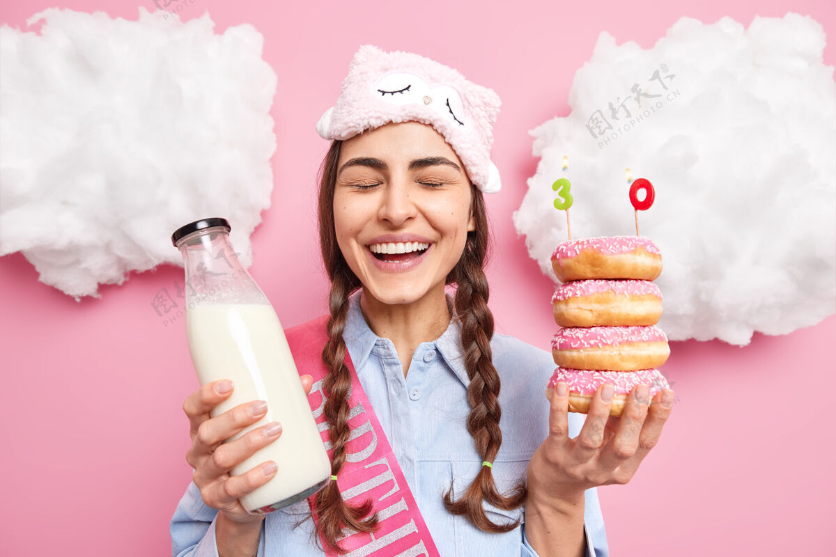 衣服女人庆祝30周年闭上眼睛笑着摆出一堆美味的甜甜圈和一瓶牛奶戴着睡衣衬衫表达幸福情绪穿模特